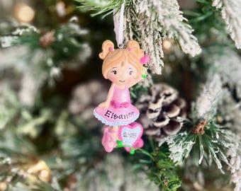 May include: A Christmas tree ornament featuring a ballerina figurine. The ornament is pink and white, with the name "Eleanor" and the year "2025" on it. The ornament is hanging on a Christmas tree with green and white branches.
