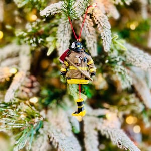 May include: A Christmas tree ornament shaped like a firefighter's uniform, including a helmet, coat, and boots. The ornament is brown, yellow, and black, with a red axe and a red string for hanging. The ornament is hanging on a Christmas tree.