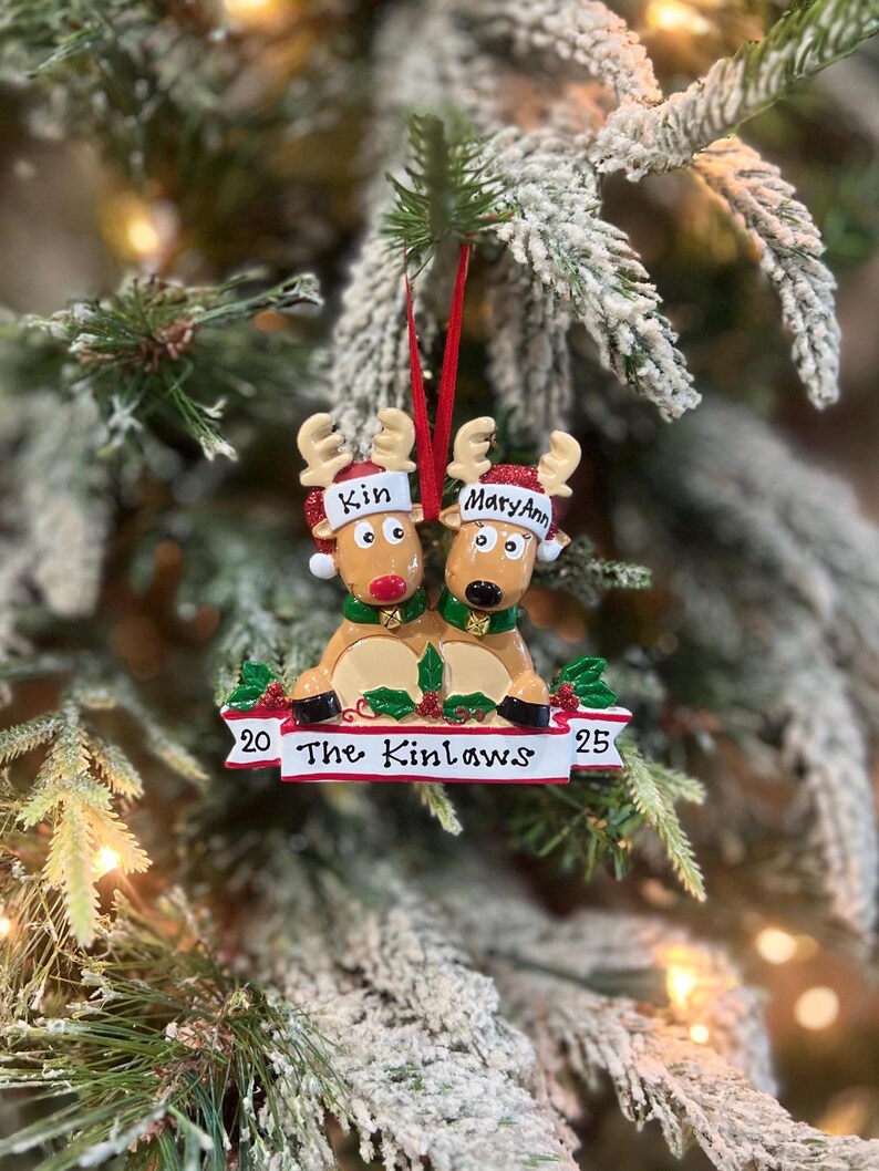 May include: A Christmas ornament featuring two reindeer wearing Santa hats and green scarves. The ornament has the names "Kin" and "MaryAnn" on the hats. The banner below reads "The Kinlaws" with the numbers "20" and "25" on either side.