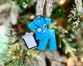 Puede incluir: Adorno navideño azul con forma de uniforme médico, con estetoscopio y portapapeles. El adorno cuelga de un árbol de Navidad con ramas verdes y blancas escarchadas.