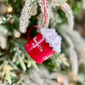 May include: A red ceramic mug Christmas ornament with white text that reads "Charlotte". The mug is filled with white whipped cream and is hanging from a red string. The ornament is hanging on a Christmas tree.