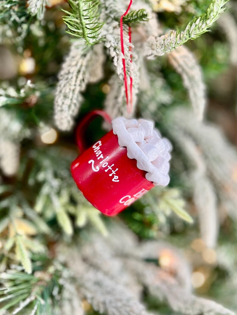 May include: A red ceramic mug Christmas ornament with white text that reads "Charlotte". The mug is filled with white whipped cream and is hanging from a red string. The ornament is hanging on a Christmas tree.