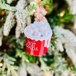 May include: A red Christmas ornament shaped like a mug of hot cocoa, with the words "HOT COCOA" in white. The ornament is topped with white marshmallow shapes and hangs from a red string. The background is a blurred Christmas tree.