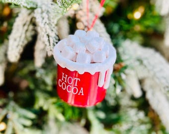 May include: A red Christmas ornament shaped like a mug of hot cocoa, with the words "HOT COCOA" in white. The ornament is topped with white marshmallow shapes and hangs from a red string. The background is a blurred Christmas tree.