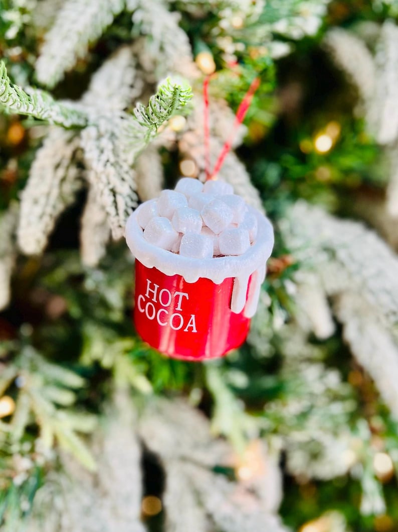May include: A red Christmas ornament shaped like a mug of hot cocoa, with the words "HOT COCOA" in white. The ornament is topped with white marshmallow shapes and hangs from a red string. The background is a blurred Christmas tree.