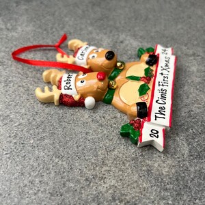 May include: A Christmas ornament featuring two reindeer with red noses and Santa hats. The ornament has a banner that reads "The Cini's First Xmas 24". The reindeer are wearing green bells and the ornament has a red ribbon for hanging.