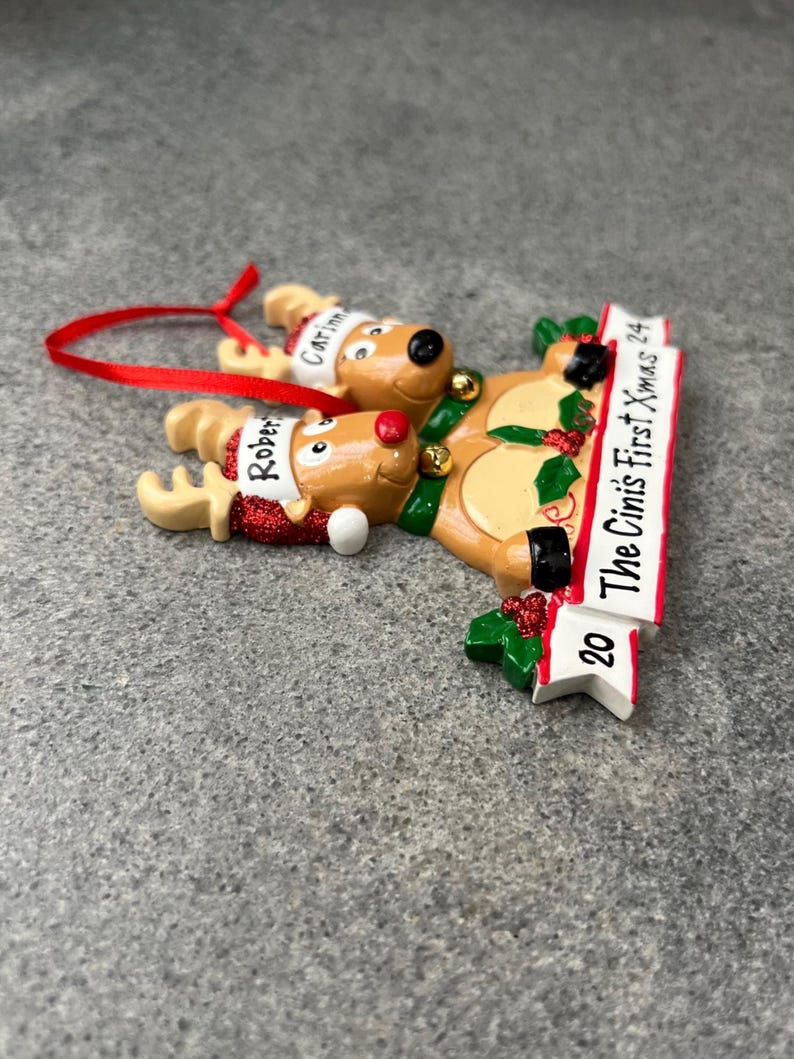 May include: A Christmas ornament featuring two reindeer with red noses and Santa hats. The ornament has a banner that reads "The Cini's First Xmas 24". The reindeer are wearing green bells and the ornament has a red ribbon for hanging.