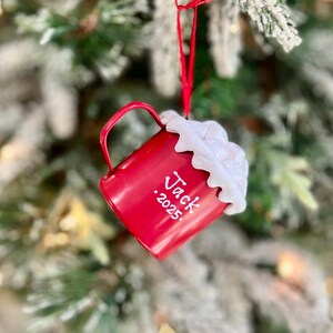 May include: A red Christmas ornament shaped like a mug, hanging from a red ribbon on a Christmas tree. The mug has white text that reads "Jack .2025" and is topped with white, fluffy material.
