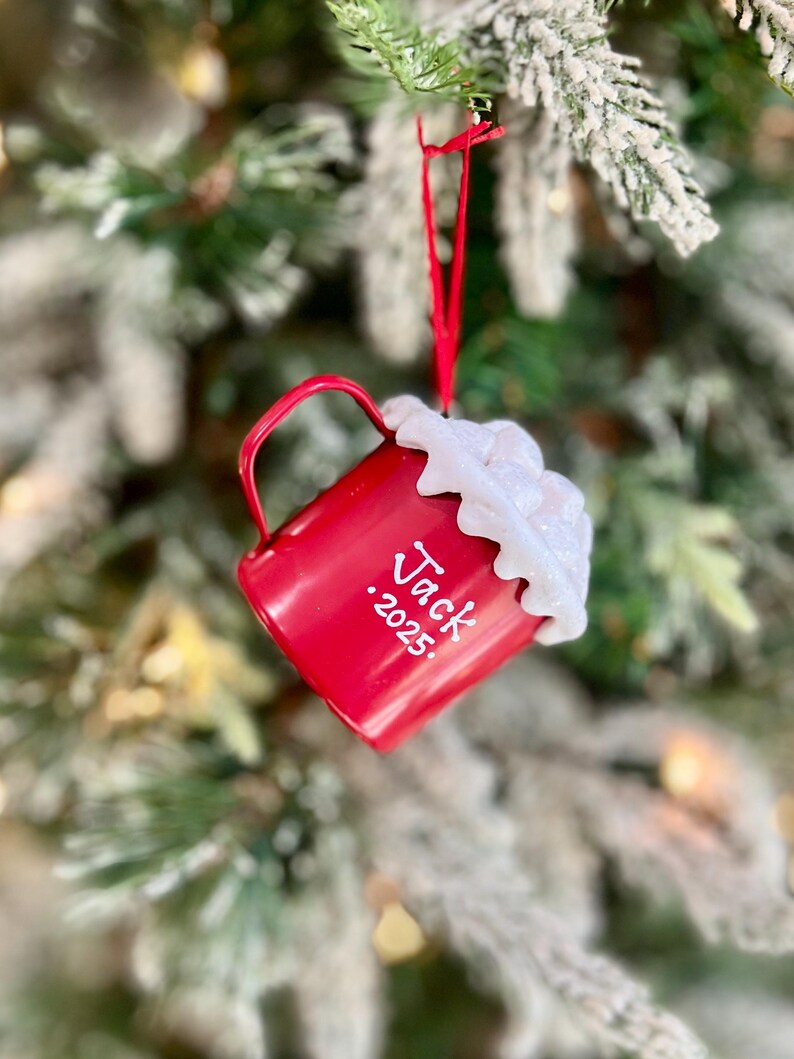 May include: A red Christmas ornament shaped like a mug, hanging from a red ribbon on a Christmas tree. The mug has white text that reads "Jack .2025" and is topped with white, fluffy material.