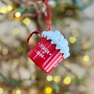 May include: A red Christmas ornament shaped like a mug, with white whipped cream detail on top. The mug has a handle and the name "Maya" and the year "2024" written on it. The ornament is hanging from a red string.
