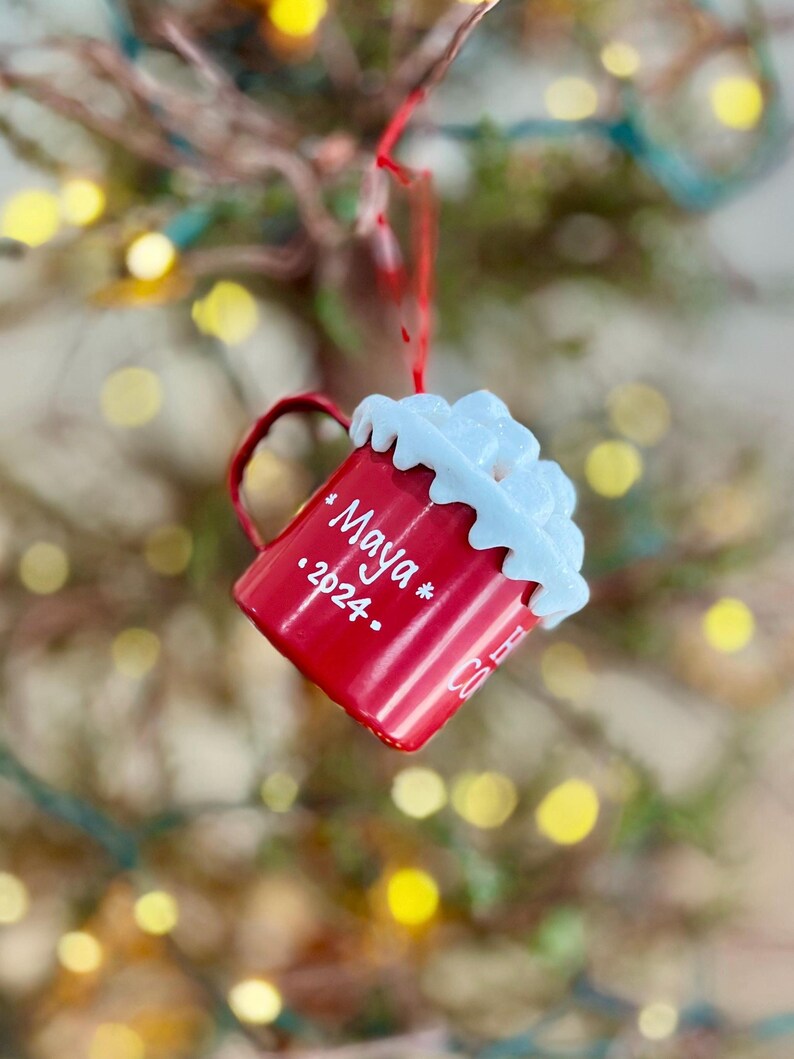 May include: A red Christmas ornament shaped like a mug, with white whipped cream detail on top. The mug has a handle and the name "Maya" and the year "2024" written on it. The ornament is hanging from a red string.