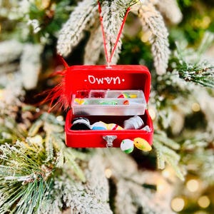 May include: A red miniature tackle box Christmas ornament with the name "Owen" written on it. The ornament is filled with tiny fishing lures and accessories. It hangs from a red string and is decorated with a red feather.