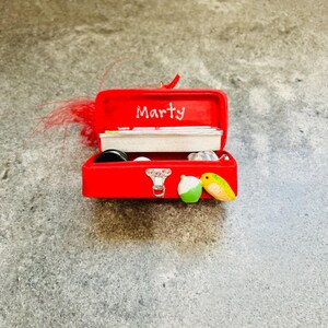 May include: A miniature red toolbox ornament with the name "Marty" in white script on the lid. The toolbox is open, revealing small fishing-themed items inside. A green and white lure and a yellow and green lure are on the front.