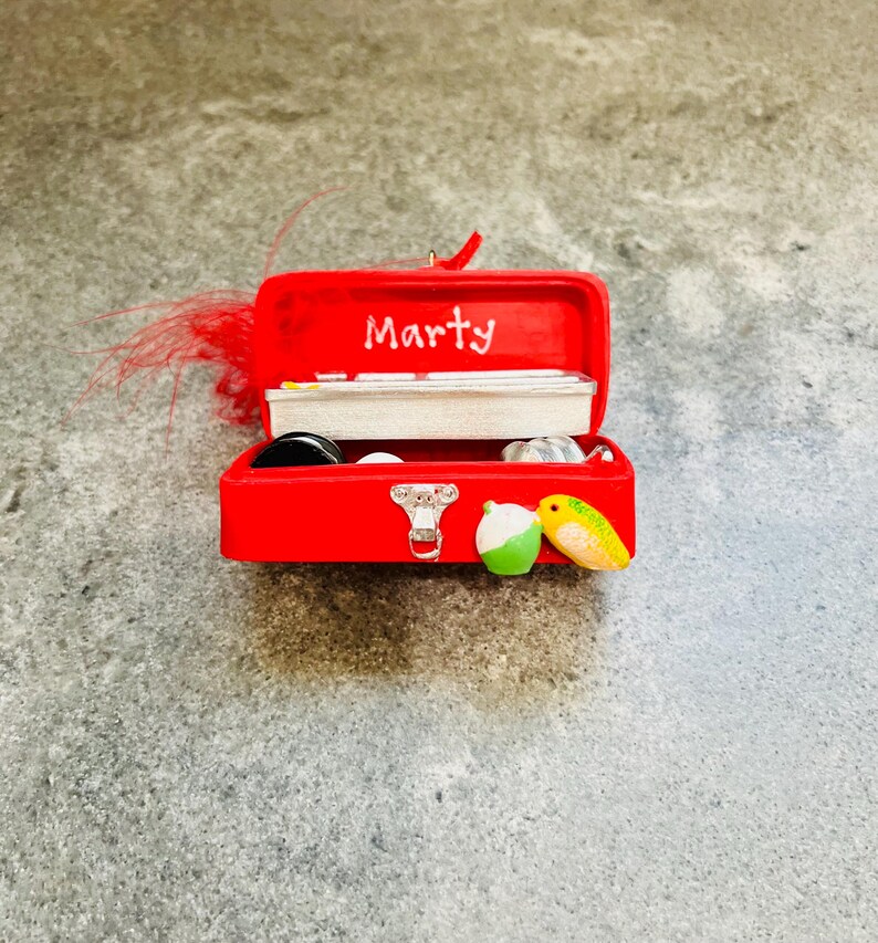 May include: A miniature red toolbox ornament with the name "Marty" in white script on the lid. The toolbox is open, revealing small fishing-themed items inside. A green and white lure and a yellow and green lure are on the front.