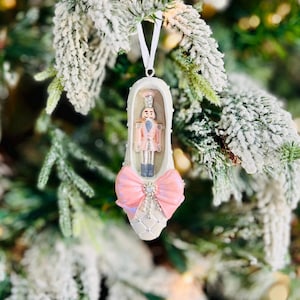 May include: A Christmas tree ornament shaped like a ballet slipper. The white slipper has a pink bow and a decorative nutcracker figure inside. The ornament hangs from a white ribbon against a backdrop of a snow-covered Christmas tree.