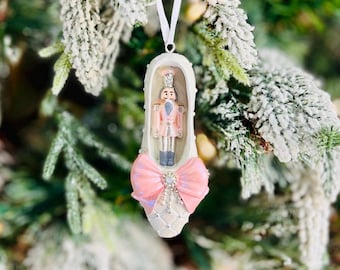 May include: A Christmas tree ornament shaped like a ballet slipper. The white slipper has a pink bow and a decorative nutcracker figure inside. The ornament hangs from a white ribbon against a backdrop of a snow-covered Christmas tree.