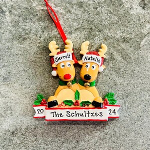 May include: A Christmas ornament featuring two reindeer with the names "Jarrett" and "Natalie" on their hats. The ornament has a banner that reads "The Schultzes 2024" and is decorated with holly and bells.