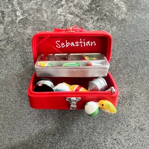 May include: A red miniature fishing tackle box with the name "Sebastian" in white script. The box is open, revealing various fishing lures and accessories in a clear plastic compartment and loose inside the box.