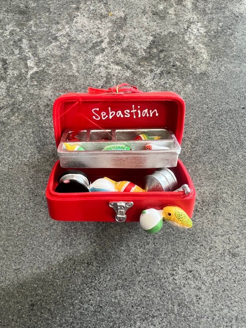 May include: A red miniature fishing tackle box with the name "Sebastian" in white script. The box is open, revealing various fishing lures and accessories in a clear plastic compartment and loose inside the box.