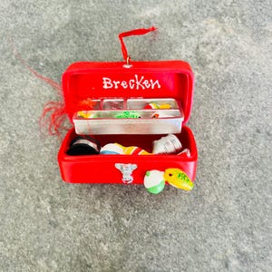 May include: A red miniature fishing tackle box ornament with the name "Brecken" in white. The box is open, revealing small fishing lures and accessories. A red loop is attached for hanging.