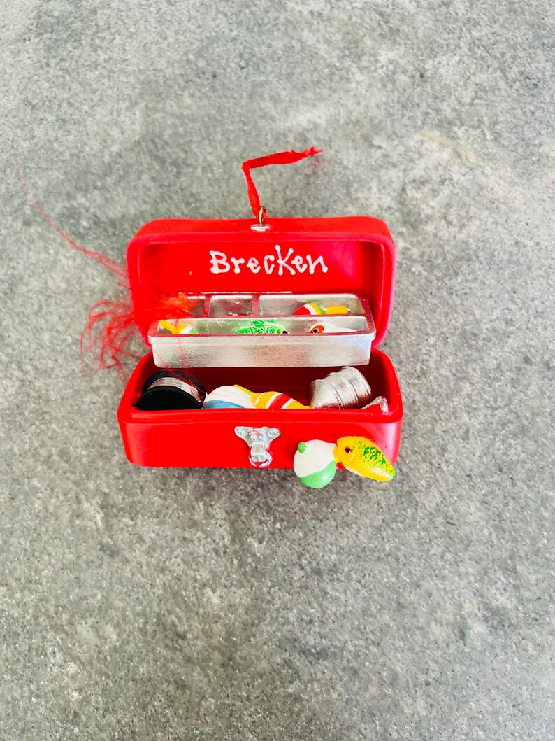 May include: A red miniature fishing tackle box ornament with the name "Brecken" in white. The box is open, revealing small fishing lures and accessories. A red loop is attached for hanging.