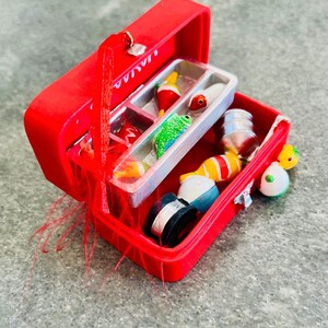 May include: A red miniature fishing tackle box, open to reveal various fishing lures, bobbers, and spools of fishing line. The tackle box is bright red with a silver clasp and handle. The contents include colorful lures and bobbers.
