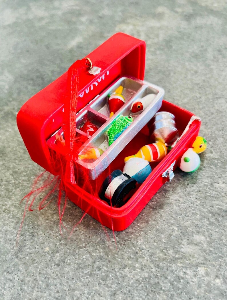 May include: A red miniature fishing tackle box, open to reveal various fishing lures, bobbers, and spools of fishing line. The tackle box is bright red with a silver clasp and handle. The contents include colorful lures and bobbers.