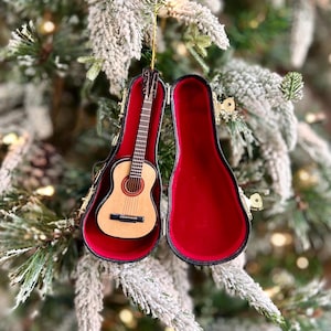 May include: A miniature acoustic guitar ornament in a black case with a red interior. The guitar is light brown with a dark brown fretboard and strings. The case is open, revealing the guitar, and is hanging from a gold string on a Christmas tree.