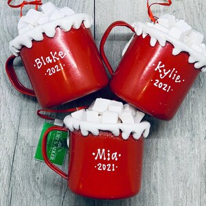 May include: Three red Christmas ornaments shaped like mugs, filled with white marshmallows and topped with a white, snowy rim. Each mug has a name and the year 2021 written in white. The mugs have red handles and are ready to hang.