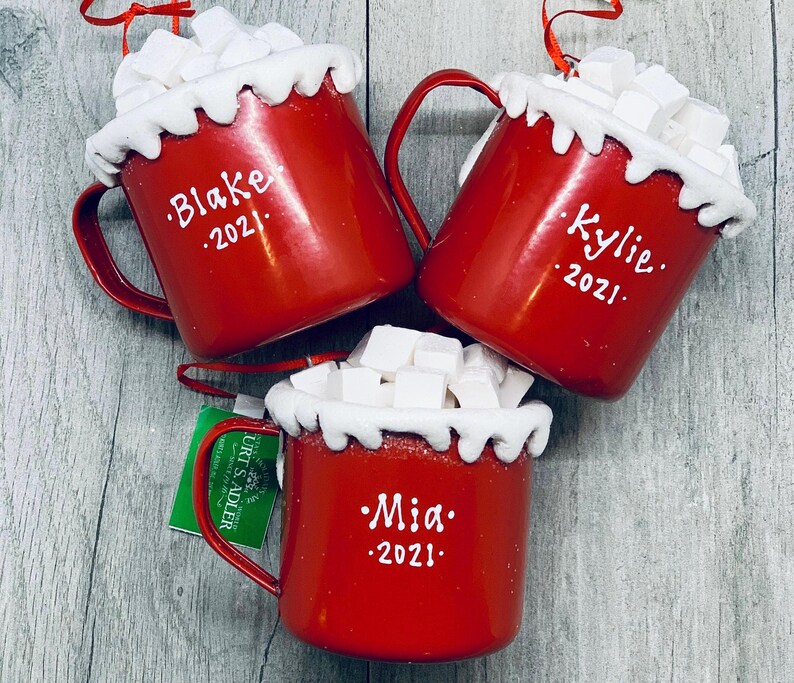 May include: Three red Christmas ornaments shaped like mugs, filled with white marshmallows and topped with a white, snowy rim. Each mug has a name and the year 2021 written in white. The mugs have red handles and are ready to hang.