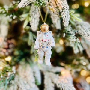 May include: A white astronaut Christmas ornament with a gold helmet and red and blue accents. The ornament is hanging from a gold string against a blurred background of a Christmas tree with snow-covered branches.
