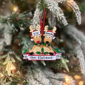 May include: A Christmas ornament featuring two reindeer with red noses and antlers. The reindeer are wearing festive hats with the names "Kin" and "MaryAnn". The ornament has a banner that reads "The Kinlaws" with the numbers "20" and "25".