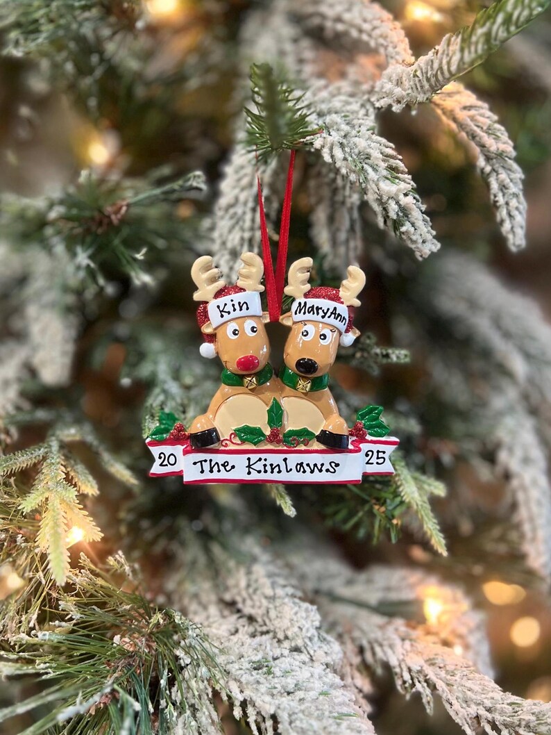 May include: A Christmas ornament featuring two reindeer with red noses and antlers. The reindeer are wearing festive hats with the names "Kin" and "MaryAnn". The ornament has a banner that reads "The Kinlaws" with the numbers "20" and "25".