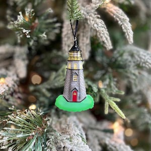 May include: A lighthouse Christmas ornament with a black top, gray brick body, and a red door. The lighthouse sits on a green base and is suspended from a black string. The ornament is hanging on a snow-covered Christmas tree.