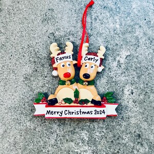 May include: A Christmas ornament featuring two reindeer with the names "Fanus" and "Carly" on their foreheads. The reindeer are tan with red noses and green collars. A banner below reads "Merry Christmas 2024".