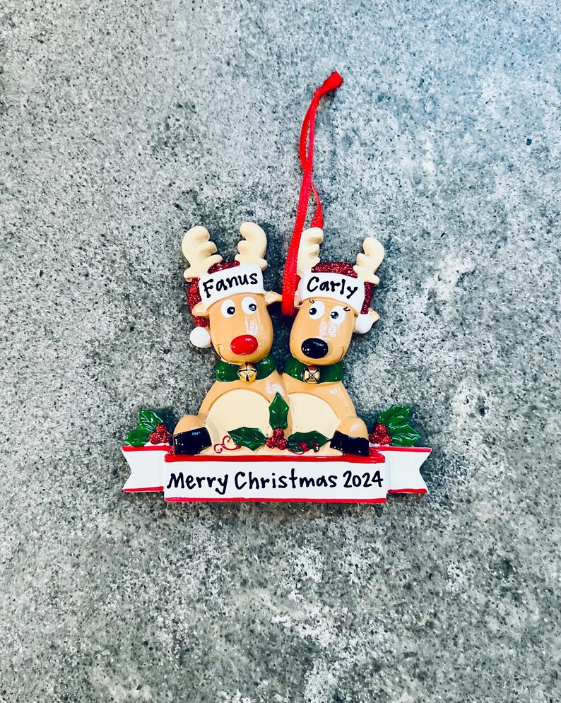 May include: A Christmas ornament featuring two reindeer with the names "Fanus" and "Carly" on their foreheads. The reindeer are tan with red noses and green collars. A banner below reads "Merry Christmas 2024".