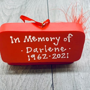 May include: A red wooden memorial ornament with the text "In Memory of Darlene 1962-2021" in white script. The ornament has a small red ribbon and a cluster of red feathers attached.