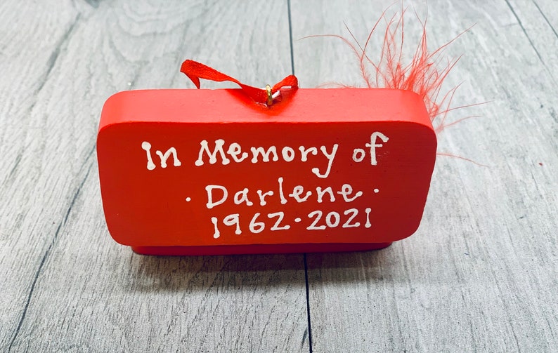 May include: A red wooden memorial ornament with the text "In Memory of Darlene 1962-2021" in white script. The ornament has a small red ribbon and a cluster of red feathers attached.