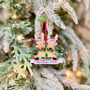 May include: A Christmas ornament featuring two reindeer with the names "Axel" and "Libby" on their hats. The reindeer are wearing green scarves and are sitting on a bed of holly. The ornament has a banner that reads "Christmas 2025!"