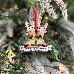 May include: A Christmas ornament featuring two reindeer with the names "Tyler" and "Amanda" on their hats. The ornament has a banner that reads "Our 4th Christmas - 1st Engaged" with the numbers "20" and "25" on either side.