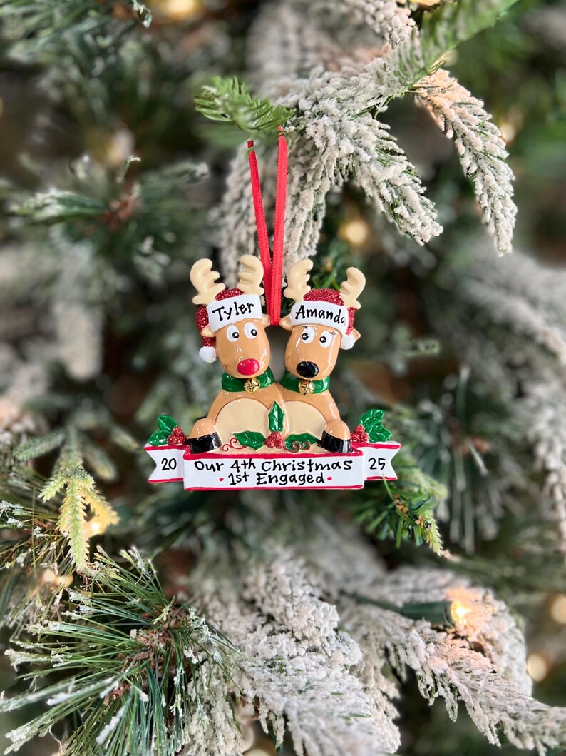 May include: A Christmas ornament featuring two reindeer with the names "Tyler" and "Amanda" on their hats. The ornament has a banner that reads "Our 4th Christmas - 1st Engaged" with the numbers "20" and "25" on either side.
