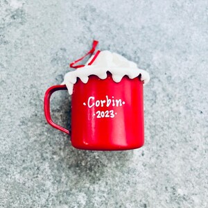 May include: A red enamel mug-shaped Christmas ornament with a white whipped cream topping. The mug has a handle and the name "Corbin" and the year "2023" in white lettering. A red string is attached for hanging.