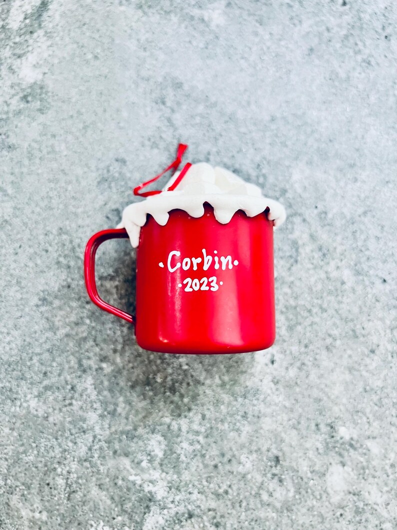 May include: A red enamel mug-shaped Christmas ornament with a white whipped cream topping. The mug has a handle and the name "Corbin" and the year "2023" in white lettering. A red string is attached for hanging.