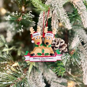 May include: A Christmas ornament featuring two reindeer wearing Santa hats with the names "George" and "Abbie". The ornament has a banner that reads "Merry Christmas 2025" and is decorated with red berries and green leaves.