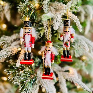 May include: Three Christmas tree ornaments shaped like nutcrackers. Each nutcracker is dressed in a red jacket, black boots, and a tall black hat. The ornaments are hanging from gold strings on a Christmas tree.