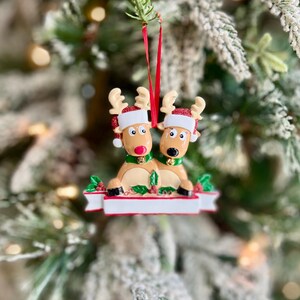 May include: A Christmas tree ornament featuring two reindeer with red and white Santa hats, green collars, and a red ribbon for hanging. The reindeer are tan with brown antlers and are positioned on a white base with red berries and green leaves.