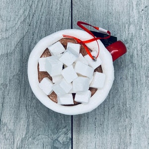 May include: A festive ornament shaped like a mug of hot chocolate, complete with white marshmallows. The mug is white with a red handle and a red ribbon for hanging. The hot chocolate is brown with white marshmallows.