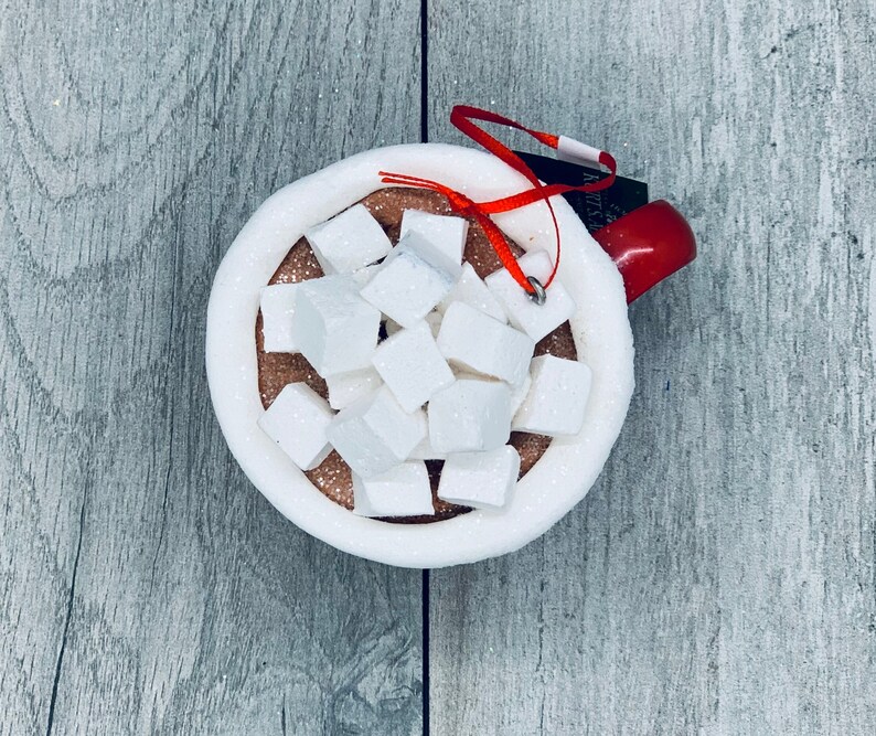 May include: A festive ornament shaped like a mug of hot chocolate, complete with white marshmallows. The mug is white with a red handle and a red ribbon for hanging. The hot chocolate is brown with white marshmallows.