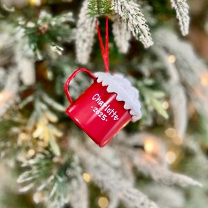 May include: A red Christmas ornament shaped like a mug, hanging from a red string on a snowy evergreen branch. The mug has a white whipped cream topping and the name "Charlotte" and the year "2025" printed on the side. The background is blurred with soft lights.