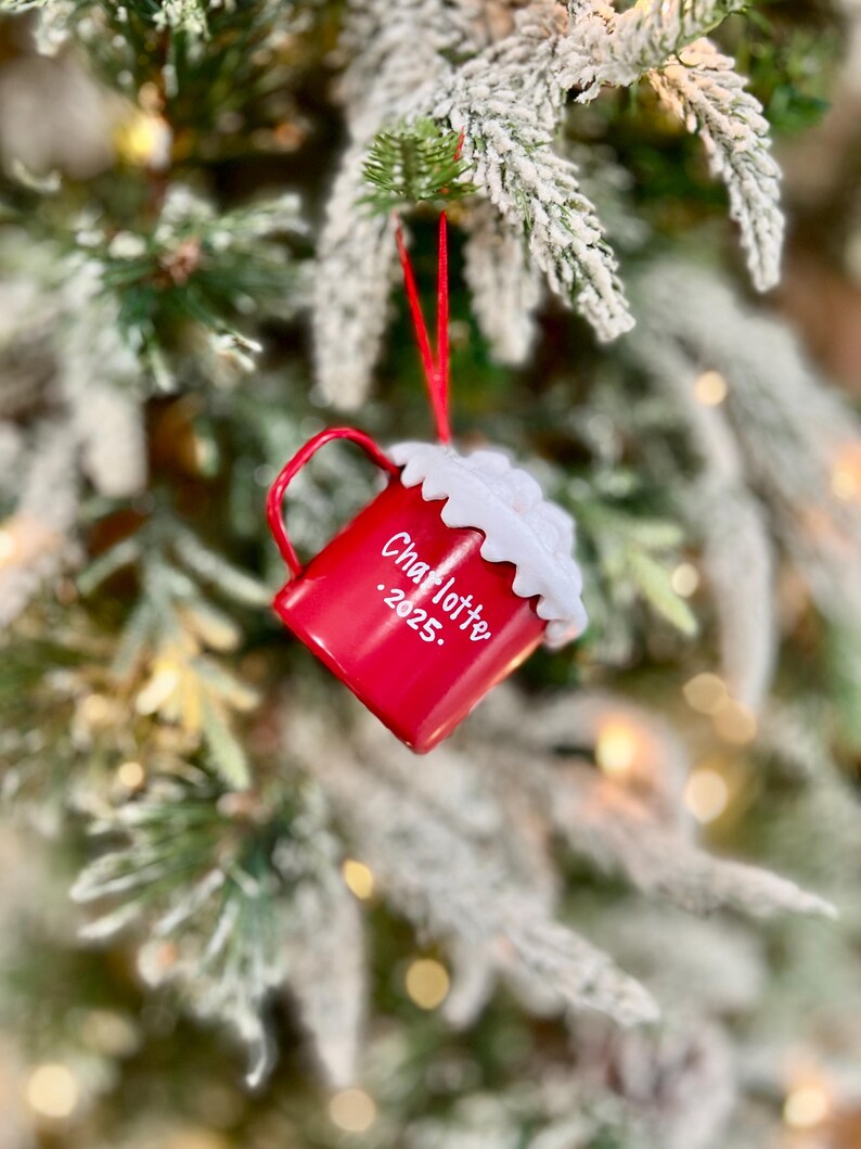 May include: A red Christmas ornament shaped like a mug, hanging from a red string on a snowy evergreen branch. The mug has a white whipped cream topping and the name "Charlotte" and the year "2025" printed on the side. The background is blurred with soft lights.